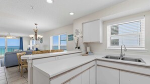 A kitchen with a white countertop, a sink, and a window with blinds. There is a dining area with a table and chairs. - Dreamy island escape with beach access, pool & complimentary seasonal beach service (Fort Walton Beach)