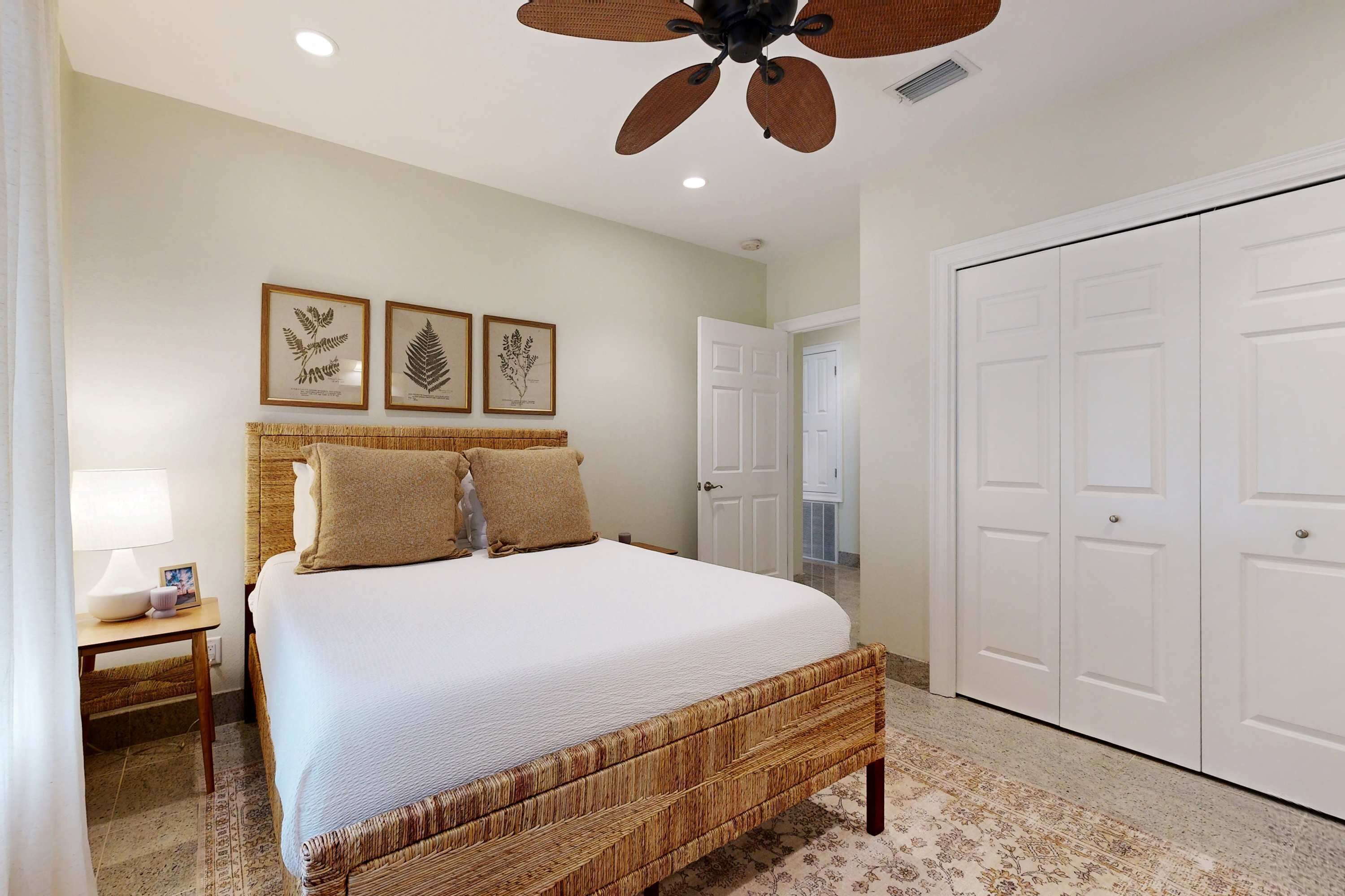 A bedroom with a large bed, a ceiling fan, a nightstand, and framed botanical prints on the wall.