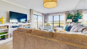 A modern living room with a large flat-screen TV, a beige sofa, and a dining area with a table and chairs. - Townhome with bay & golf views, golf cart, pool, & beach tram (Miramar Beach)