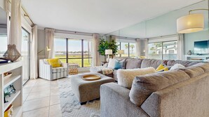 A modern living room with a sectional sofa, ottomans, and a large window offering a view of the outdoors. - Townhome with bay & golf views, golf cart, pool, & beach tram (Miramar Beach)
