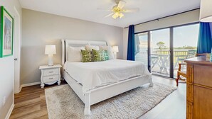 Un dormitorio con cama, mesitas de noche, ventilador de techo, ventana grande y vistas al exterior.