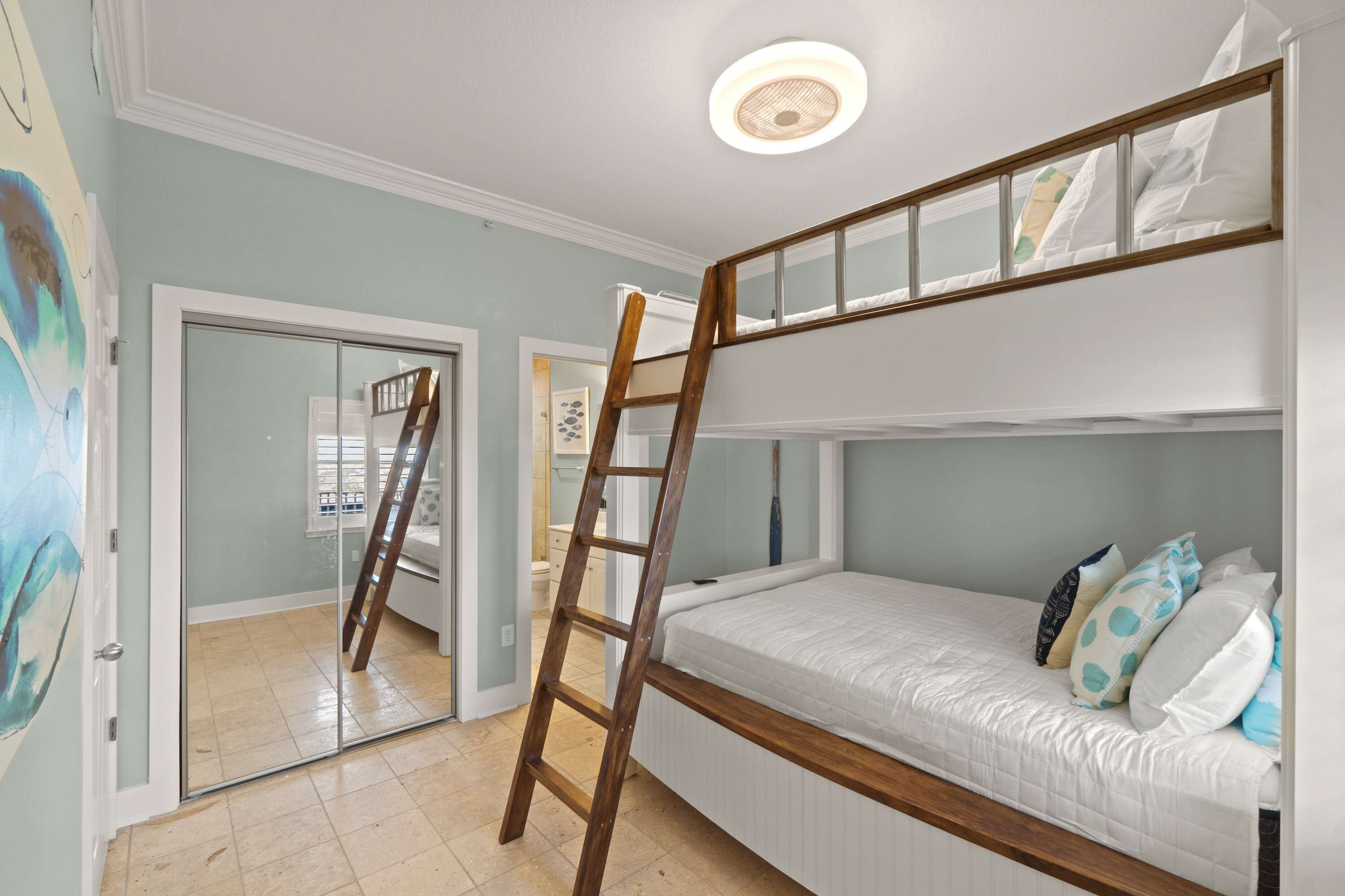 Habitación con literas, una escalera de madera, una lámpara en el techo y una puerta que da a otra habitación.