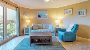 Un salon comprenant un canapé, un fauteuil, une table basse et deux petites tables d’appoint avec des lampes. On y trouve également un ventilateur de plafond, une grande fenêtre avec des rideaux et des tableaux encadrés accrochés au mur.