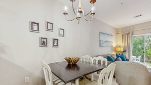 Un coin repas avec une table en bois foncé, des chaises blanches et un lustre. Des tableaux encadrés ornent le mur, et une fenêtre est agrémentée de rideaux.