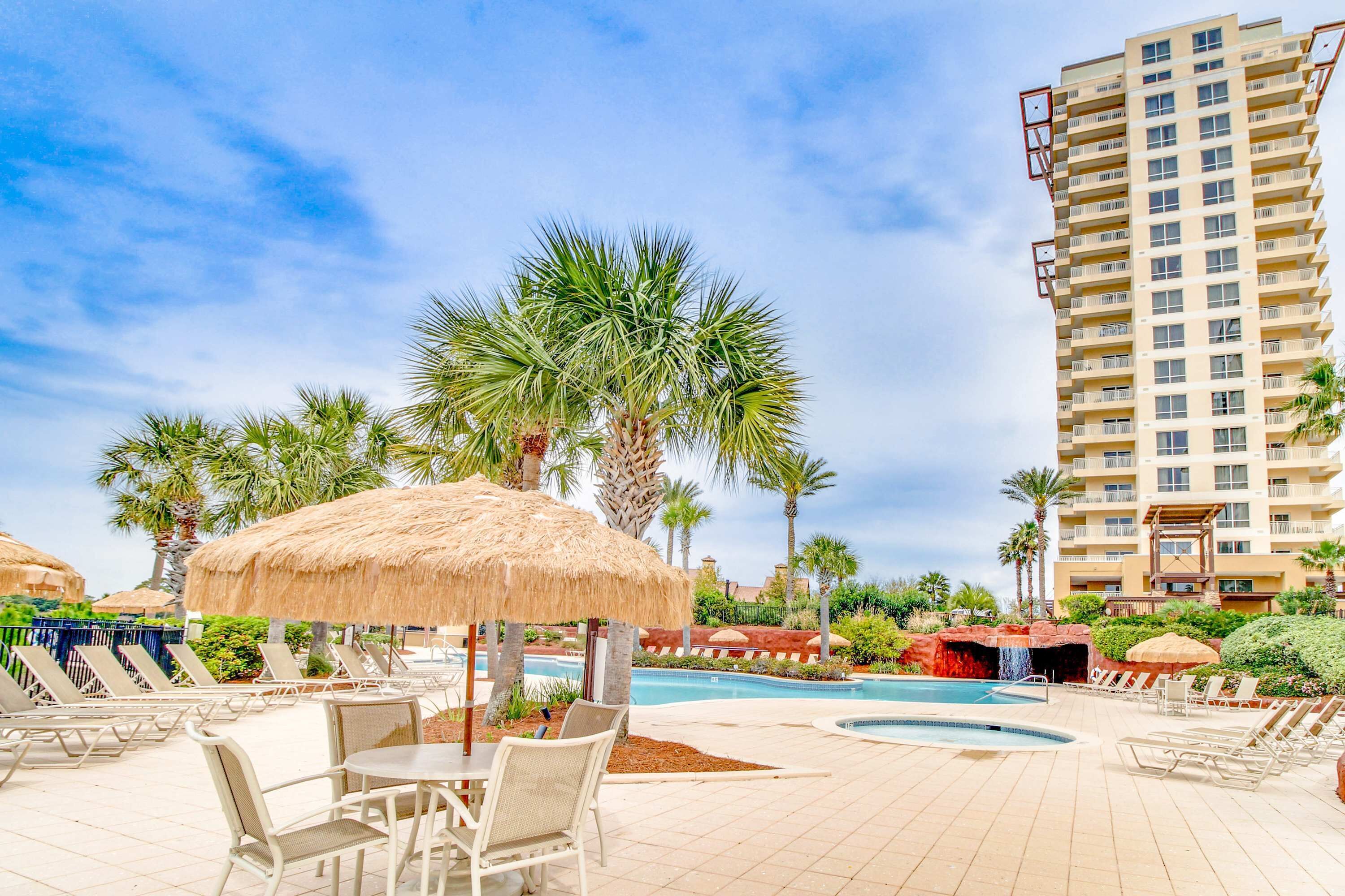 A resort pool area with lounge chairs, umbrellas, and a tall building in the background.