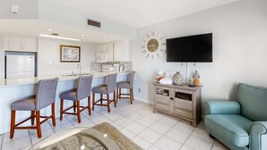 A modern kitchen with a central island, bar stools, a large wall clock, a flat-screen TV, and a teal sofa in a living area. - Large waterfront cottage, Private pool, Close to entertainment (Miramar Beach)