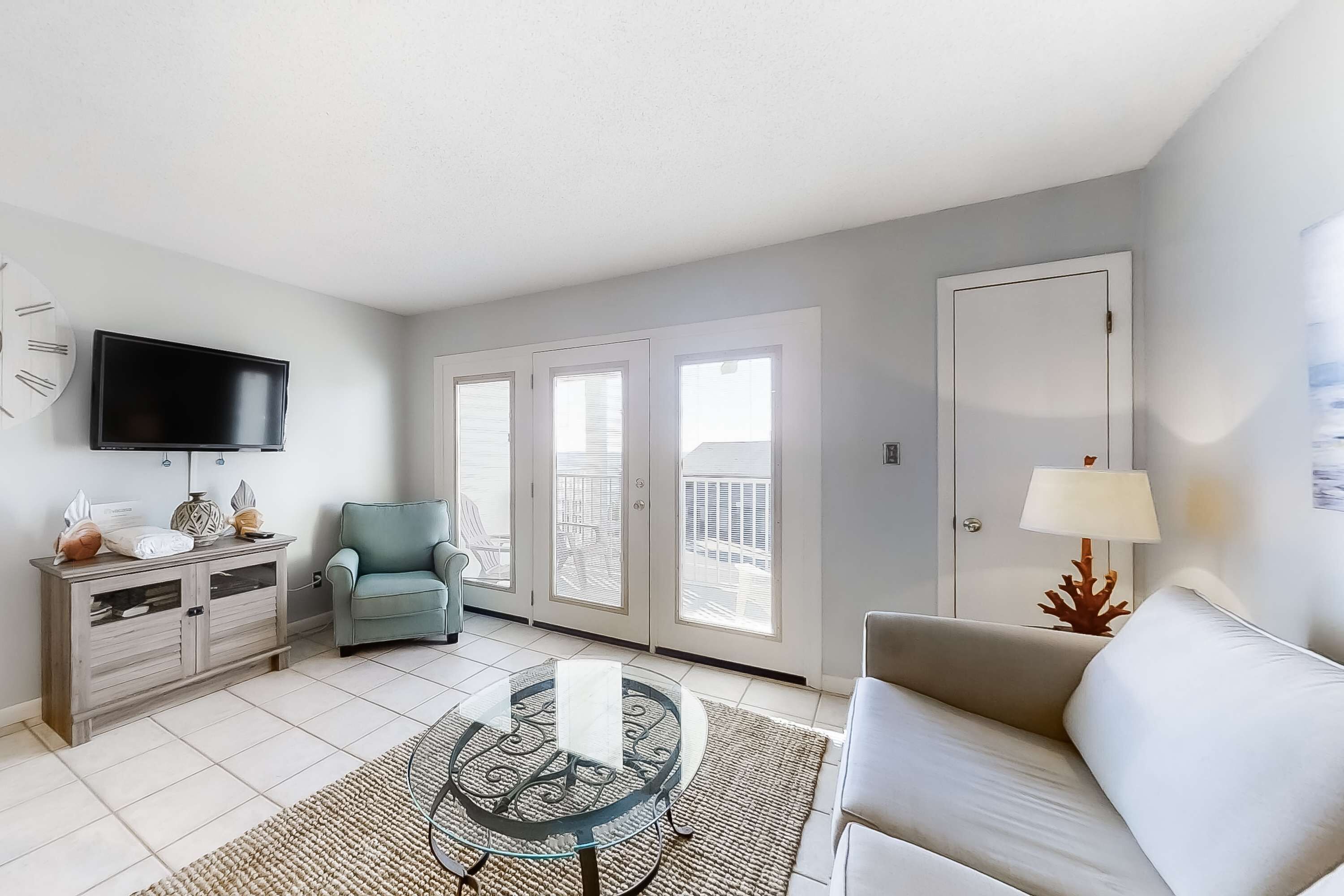 Un salon comprenant un canapé, une table basse avec un plateau en verre, un téléviseur, un fauteuil et une porte donnant sur un balcon.