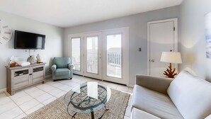 A living room with a sofa, a glass-top coffee table, a TV, a chair, and a door leading to a balcony. - Large waterfront cottage, Private pool, Close to entertainment (Miramar Beach)