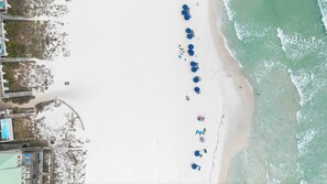 Vista aérea de uma praia com uma fileira de guarda-sóis azuis e espreguiçadeiras, um calçadão de madeira e um prédio com piscina.