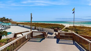 Eine Holzterrasse führt zu einem Strand mit einer Flagge, einem Pool und Liegestühlen.