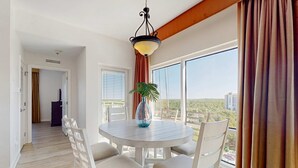 A dining area with a round table, white chairs, a hanging light fixture, and a view of the city through large windows. - Luau getaway with pools, hot tub, beach access, & private balcony (Miramar Beach)
