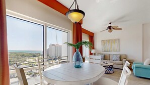 A modern living room with a round dining table, a sofa, a ceiling fan, and a large window offering a city view. - Luau getaway with pools, hot tub, beach access, & private balcony (Miramar Beach)