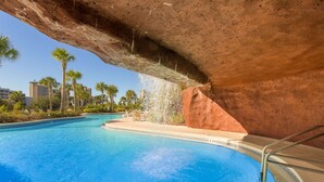 A swimming pool with a waterfall feature, surrounded by palm trees and a red rock structure. - Luau getaway with pools, hot tub, beach access, & private balcony (Miramar Beach)