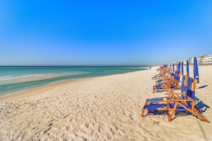 Ein Strand mit Reihen von blauen und orangefarbenen Liegestühlen und Sonnenschirmen, einem Sandstrand und einem klaren, blauen Himmel.