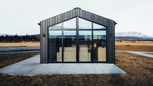A modern, single-story building with large glass windows and a metal roof, situated in a rural landscape with mountains in the background. - Green Geysir Cottage (Bláskógabyggð)