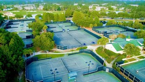 Eine Vogelperspektive zeigt einen Sportkomplex mit mehreren Tennisplätzen, umgeben von Bäumen und Gebäuden.