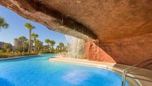 Une piscine agrémentée d’une cascade, entourée de palmiers et surplombée d’un ciel dégagé.