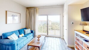 Un salon moderne avec un canapé bleu, une table basse en bois et une grande fenêtre donnant sur un balcon et des immeubles.
