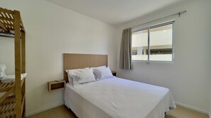 Un lit simple avec une tête de lit en bois, une table de chevet, une fenêtre avec des rideaux et une étagère en bois où sont pliés des vêtements.