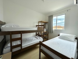 Quarto com beliches, prateleira de madeira, janela com cortinas e vista para o exterior.