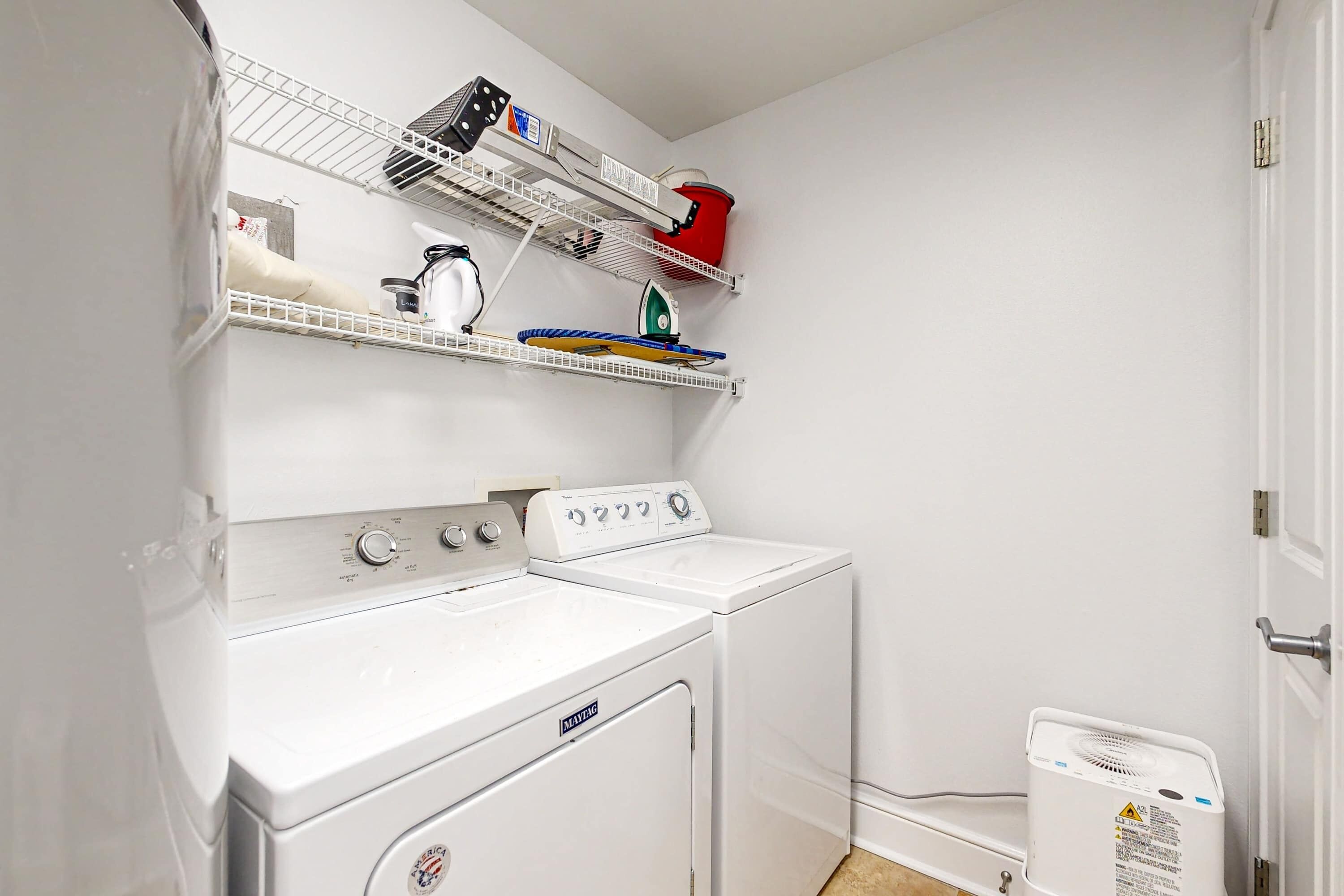 A laundry area with a front-loading washer and dryer, a shelf with various items, and a small heater on the floor.