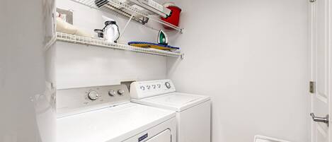 A laundry area with a front-loading washer and dryer, a shelf with various items, and a small heater on the floor.