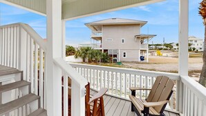 A covered porch with wooden steps and white railings, overlooking a beach with a two-story building in the background. - Spacious home with private pool & full kitchen, close to beach (St. George Island)