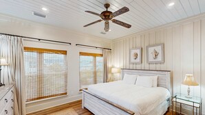 A bedroom with a wooden bed, a ceiling fan, two windows with blinds, a bedside table with a lamp, and a dresser. - Spacious home with private pool & full kitchen, close to beach (St. George Island)