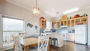 A modern kitchen with wooden cabinets, a central island, and a dining area with white chairs. - Beachfront dog-friendly home with private hot tub, boardwalk, on-site pools, gym (St. George Island)