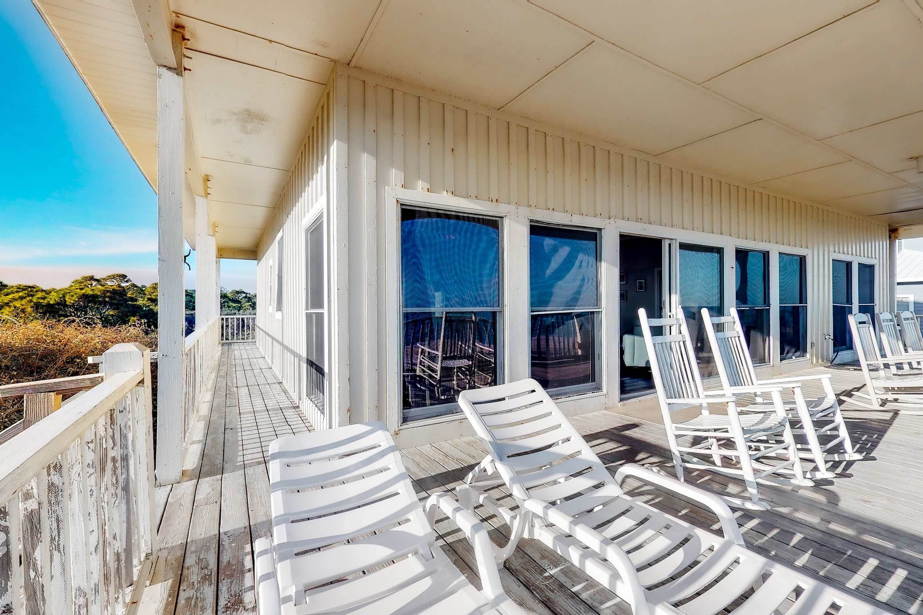 A covered deck with white lounge chairs and a wooden floor, overlooking a beach with vegetation.