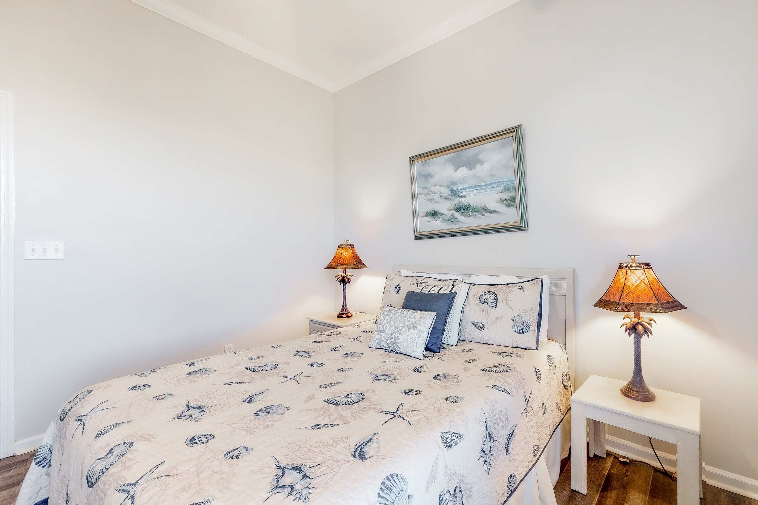 A bedroom with a bed, two bedside tables, two lamps, a painting on the wall, and a patterned bedspread.