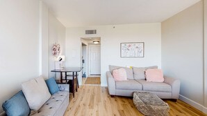 Ein Wohnzimmer mit grauem Sofa, einem kleineren grauen Sofa, Holzboden, einem kleinen Tisch mit Lampe und einem gerahmten Bild an der Wand.