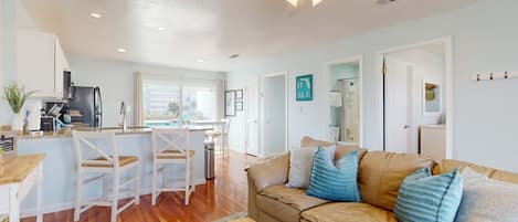 A spacious living area with a beige sofa, a wooden coffee table, and a kitchen area with white appliances and bar stools.