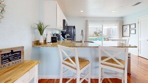 Una cocina moderna con armarios blancos, encimera de granito y vistas a la piscina. Hay dos sillas blancas con asientos de mimbre.