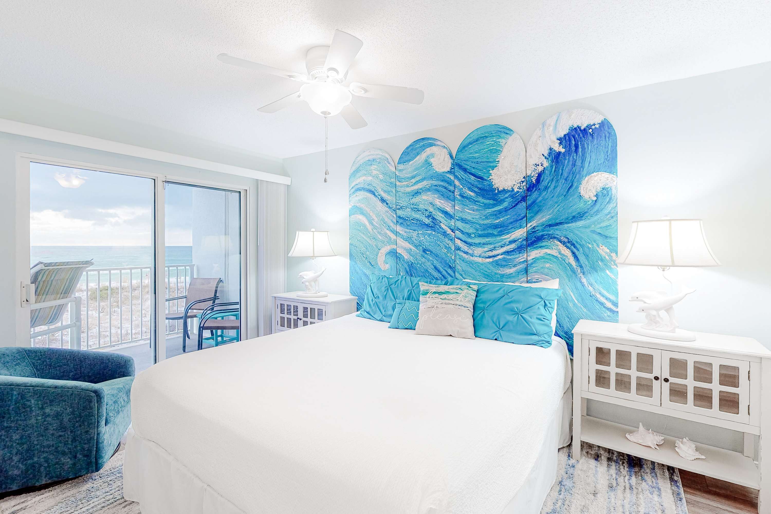 Een hotelkamer met een groot bed, een plafondventilator, uitzicht op zee, een balkon met stoelen en een groot schilderij van een blauwe golf aan de muur.