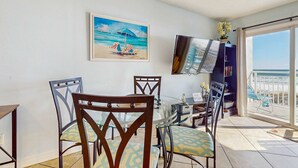 Un comedor con mesa y sillas de vidrio, un cuadro con escena de playa, un televisor montado en la pared y un balcón con vista al océano.