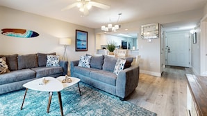 A living room with a grey sofa, a white coffee table, and a blue rug. There is a ceiling fan, a chandelier, and a surfboard decoration on the wall. - Gorgeous coastal home with plenty of space and easy beach access (Fort Walton Beach)