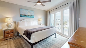 A hotel room with a large bed, a ceiling fan, a painting of sailboats, two bedside lamps, and a view of a balcony. - Gorgeous coastal home with plenty of space and easy beach access (Fort Walton Beach)