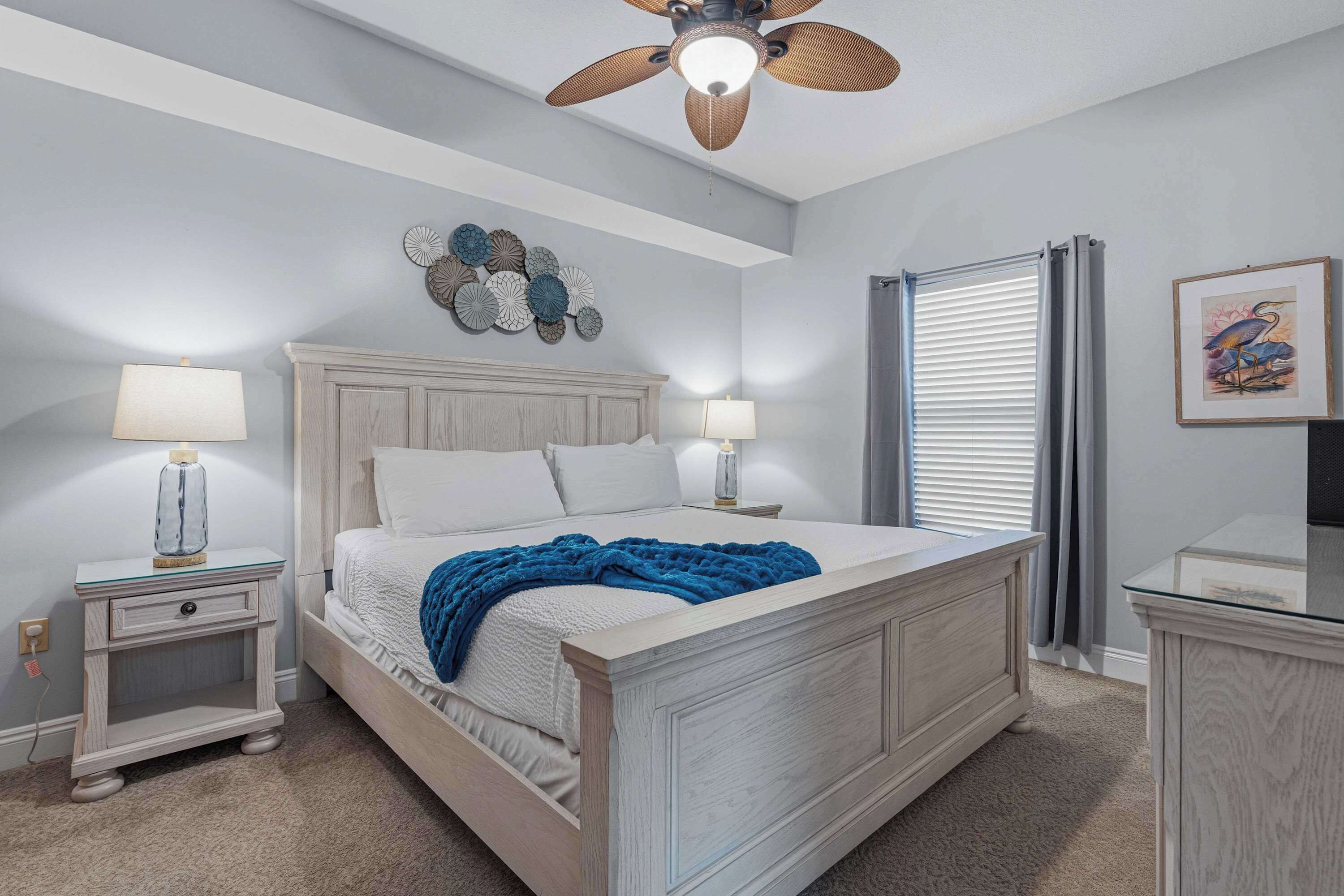 A bedroom with a bed, two bedside tables, a ceiling fan, a wall clock, a decorative wall piece, a window with curtains, and a framed picture on the wall.
