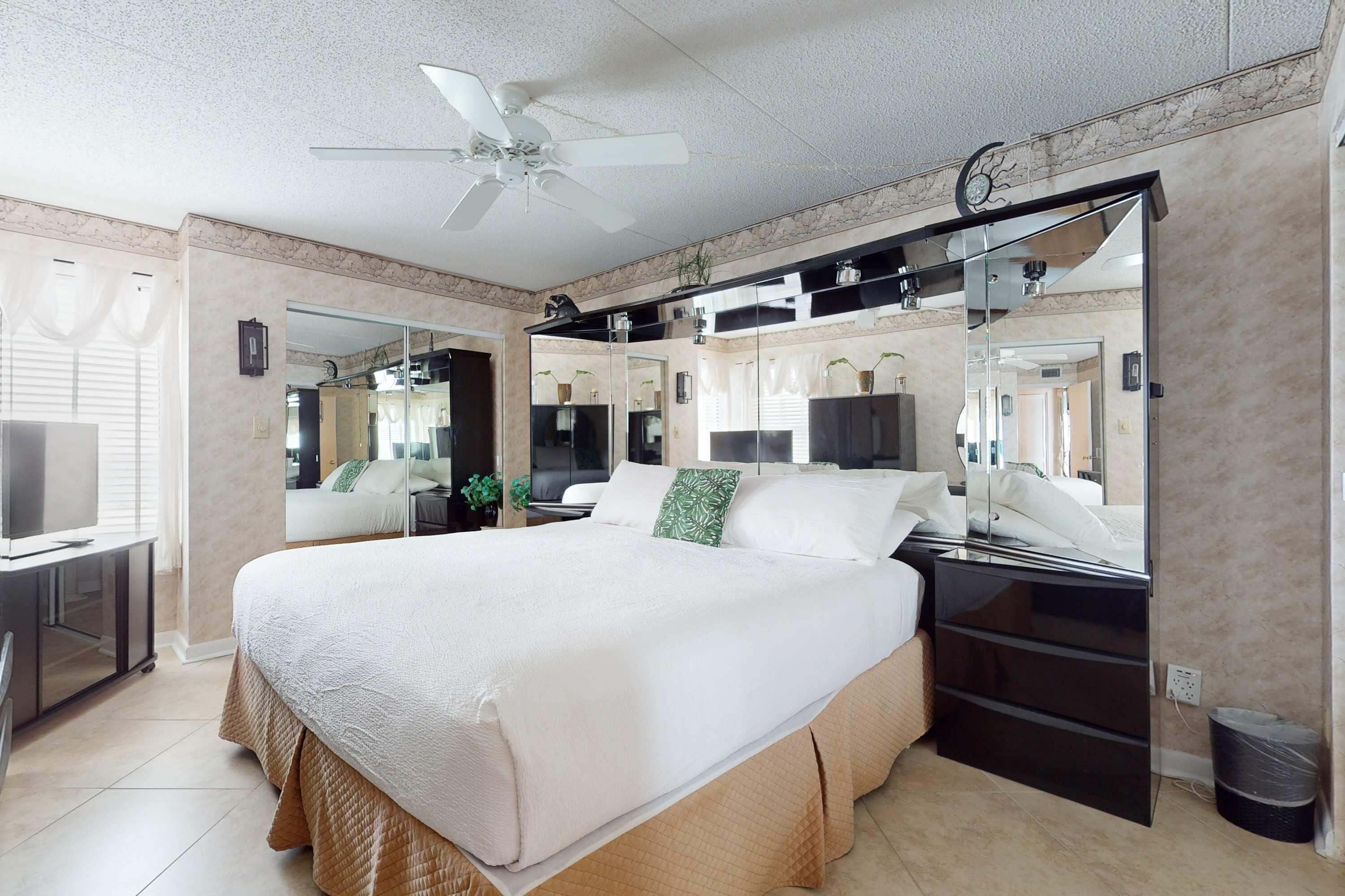 Ein geräumiges Schlafzimmer mit einem großen Bett, einem Deckenventilator, einem Fernseher und einem Spiegel.