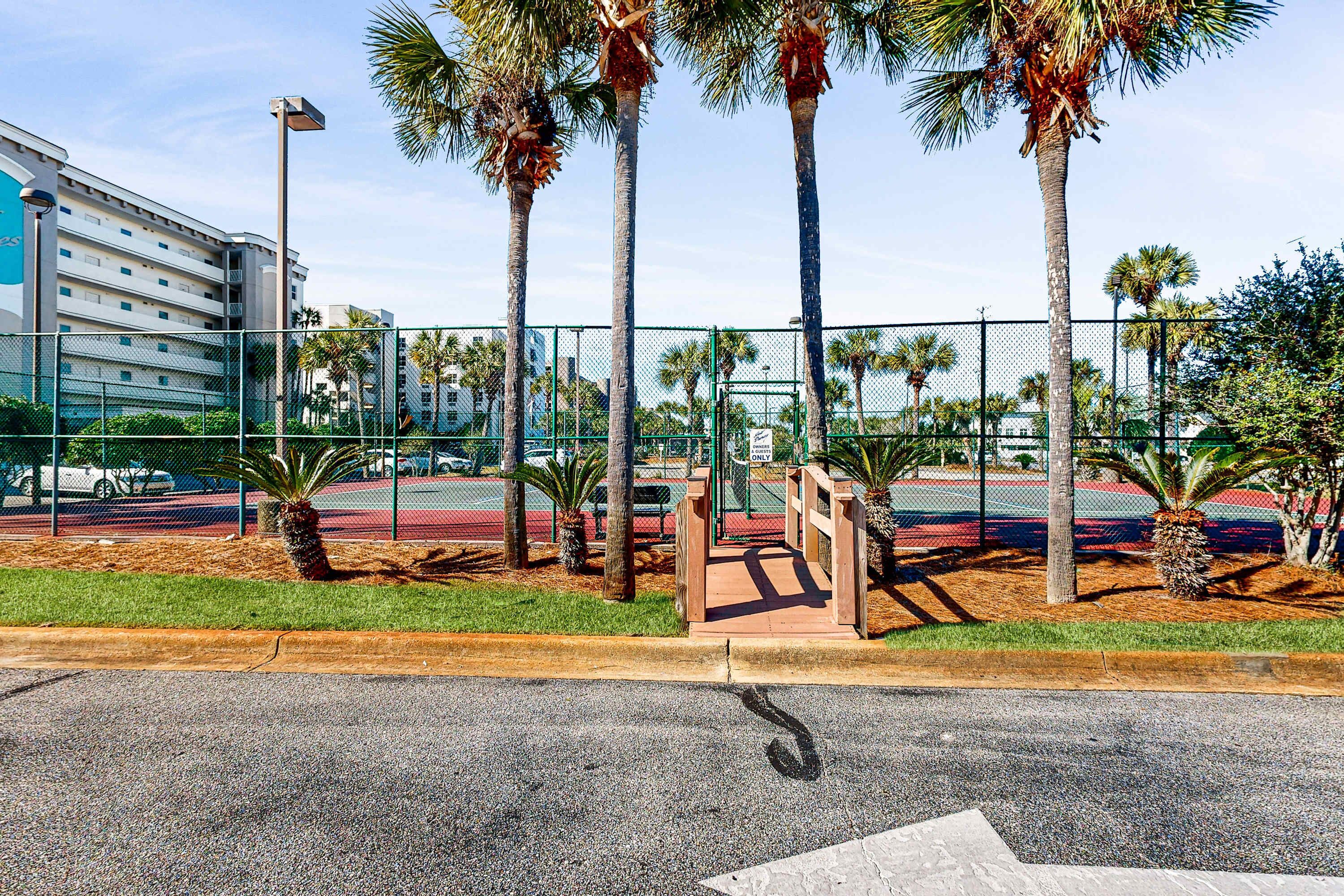 A fenced tennis court area with palm trees and a building in the background.