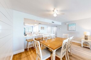A dining area with a wooden table and white chairs, a ceiling fan, and a painting on the wall. - Gulf-front condo with HD flat-screen televisions Tiki bar & Sky bar on-site (Miramar Beach)