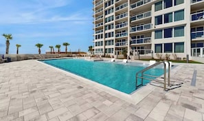 Une piscine dont le fond est recouvert de carreaux disposés en motif de chevron, une échelle de piscine en métal et, au fond, un immeuble à plusieurs étages doté de balcons.