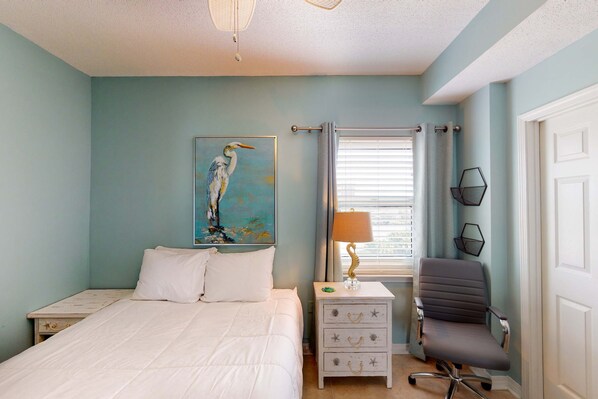 A bedroom with a bed, a nightstand, a desk, a chair, a window with blinds, and a painting of a bird. - 4th Floor Condo, On-Site Pool, Amazing View, Beach-Front (Pensacola Beach)