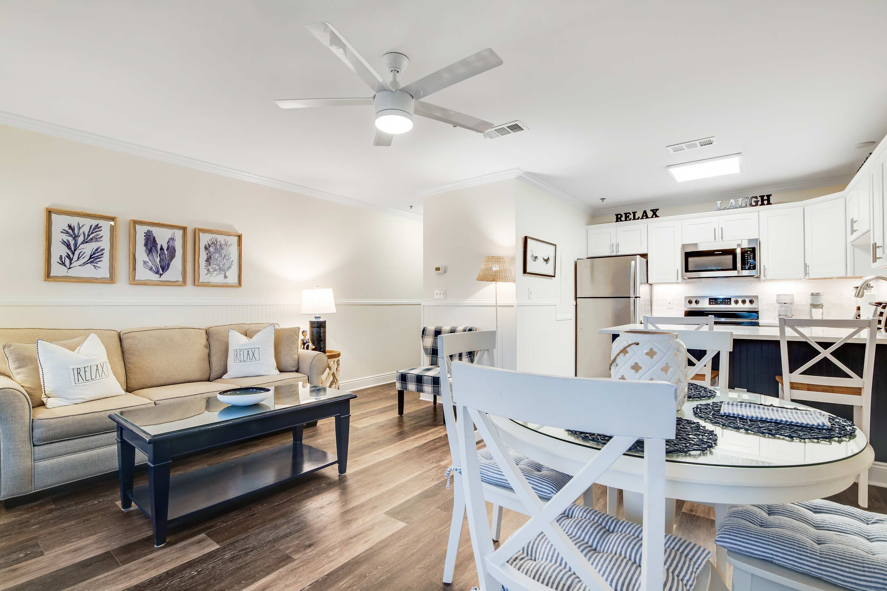 Un salon moderne comprenant un canapé, une table basse et des fauteuils, un coin repas avec une table et des chaises, et une cuisine dotée de meubles blancs et d’appareils électroménagers.
