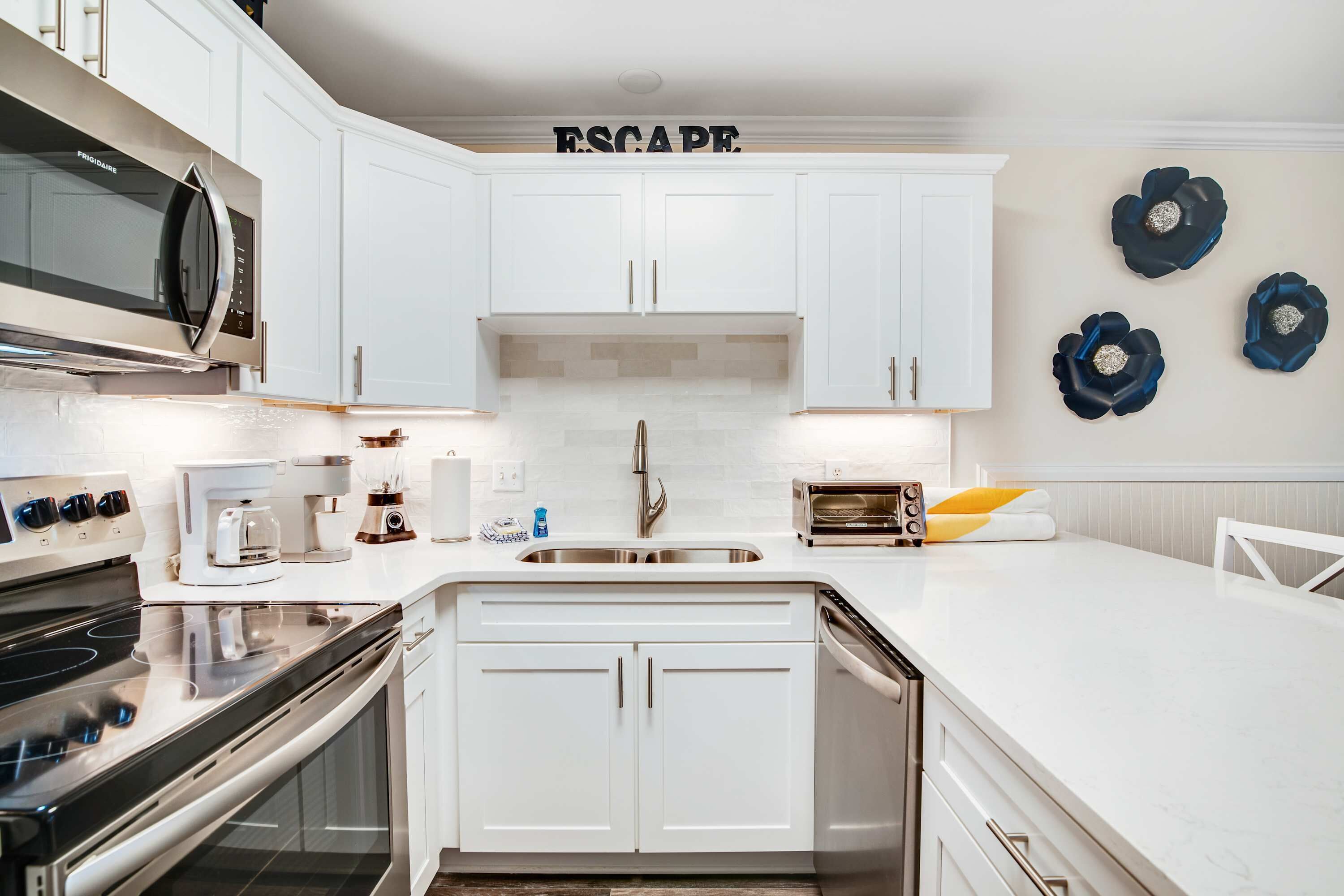 A modern kitchen with white cabinetry, a stainless steel microwave, a built-in oven, and a sink. There are decorative wall art pieces and a word "ESCAPE" on the wall.