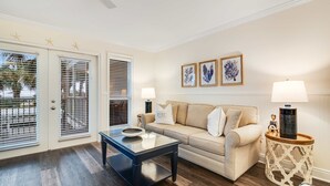 Een woonkamer met een beige bank, een glazen salontafel en een bijzettafeltje met een lamp. Drie ingelijste foto's hangen aan de muur en aan het plafond zien we decoraties van zeesterren.