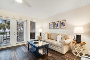 A living room with a beige sofa, a glass coffee table, and a side table with a lamp. There are three framed pictures on the wall and starfish decorations on the ceiling. - Charming condo near the beach, with community pools and hot tub (Santa Rosa Beach)
