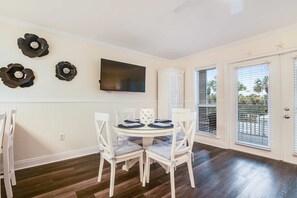 A dining area with a round table, white chairs, and a flat-screen TV mounted on the wall. - Charming condo near the beach, with community pools and hot tub (Santa Rosa Beach)