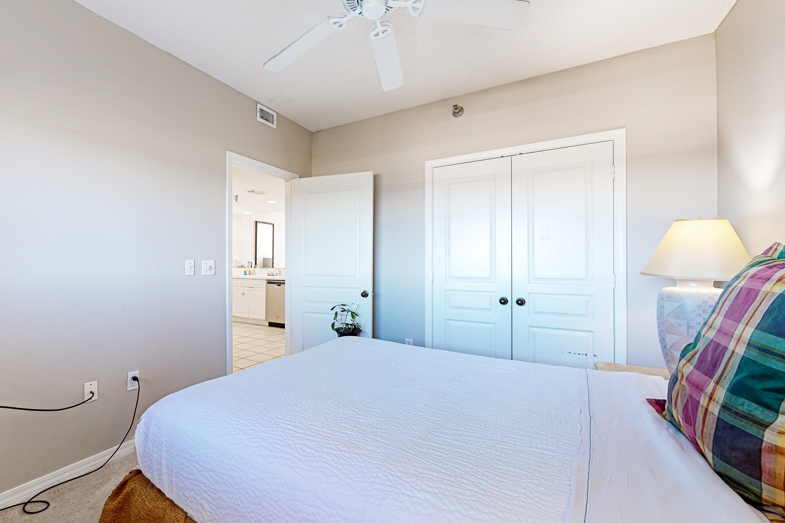 A bedroom with a bed, a ceiling fan, a lamp, and a door leading to a bathroom.